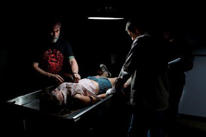 People examining a body on a metal table under a single overhead light in a dark room