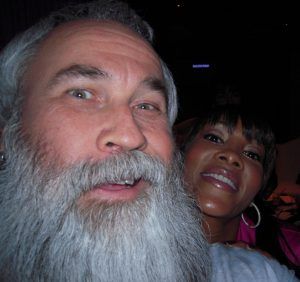 Close-up selfie of two smiling people in a dark indoor setting, one with a long gray beard