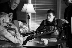 Two men in a dim room aim a rifle across a table beside a lit lamp and cup.