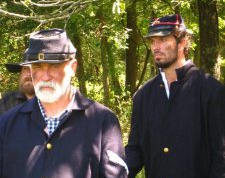 Two men in dark coats and hats standing outdoors in a sunlit wooded area