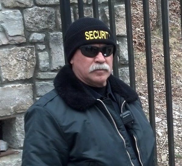 Security guard in black beanie and sunglasses standing by a stone wall and iron gate
