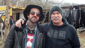 Two men posing outdoors, one in a black hat and sunglasses with his arm around the other.