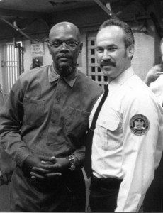 Two men standing together indoors, one in a dark shirt and the other in a white uniform shirt with a badge patch.
