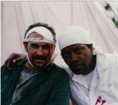 Two men with head bandages and blood-stained clothes pose closely in front of a light tent backdrop