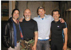 Four men smiling and standing close together indoors, wearing casual dark clothing.