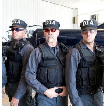 Three DIA agents in black tactical vests and caps stand together indoors, wearing sunglasses.