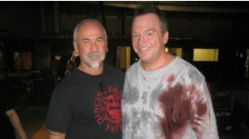 Two smiling men pose indoors, one in a black shirt, the other in a gray tie-dye shirt with a red stain.