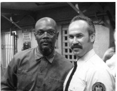 Two men standing together indoors, one in a dark shirt and one in a light shirt, both facing the camera.