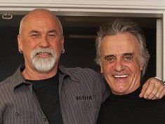 Two smiling men standing together indoors, one with an arm around the other