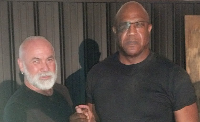 Two men standing indoors, one holding up a small object, against a dark curtain backdrop.