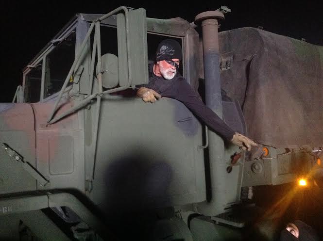 Person leaning out of a large military truck at night, wearing a face covering and pointing forward.