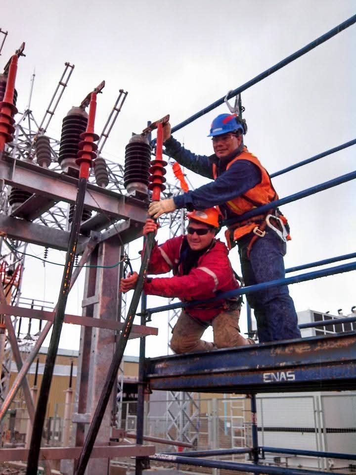 Dos trabajadores con equipo de seguridad escalando una torre de transmisión eléctrica en una subestación al aire libre.
