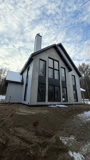 Modern gray house with black-framed windows and chimney under a cloudy sky, snow on the ground.
