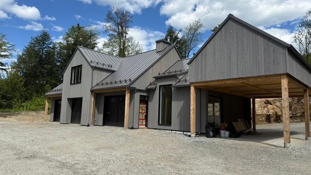 Maison grise moderne avec abri voiture, toit en métal et grandes fenêtres sur un terrain gravillonné.