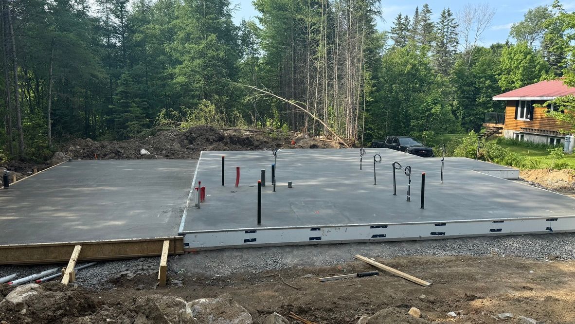 Fondations en béton avec canalisations, devant des arbres et une maison.