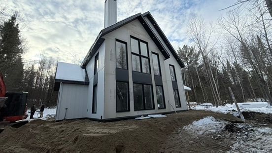 Modern white house with dark trim and tall windows, chimney, set on a snowy hill, surrounded by trees.