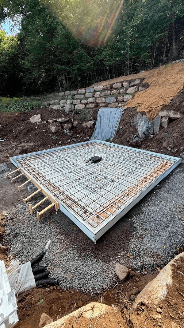 Construction site: Square concrete foundation with rock edging, set into a graded hillside.