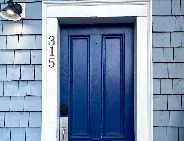 Blue door with white trim and house number 315 on a blue-shingled exterior, illuminated by a black light fixture.