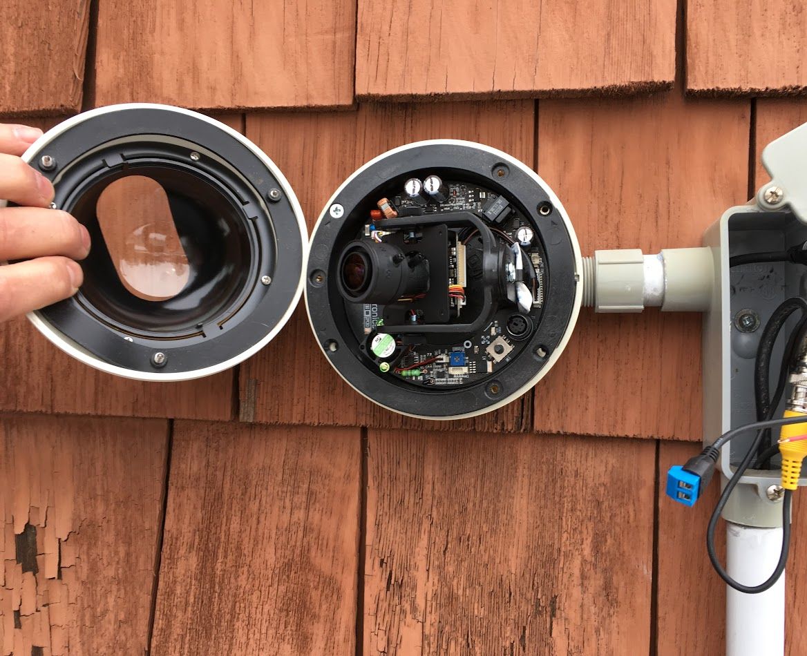 Outdoor security camera, opened for repair, mounted on a wooden wall.