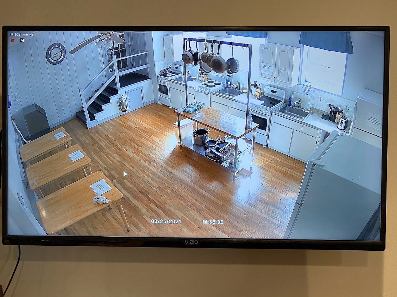 Monitor displays an overhead view of a kitchen. Wooden floors, stainless steel cooking equipment, white cabinets, tables, and staircase are visible.