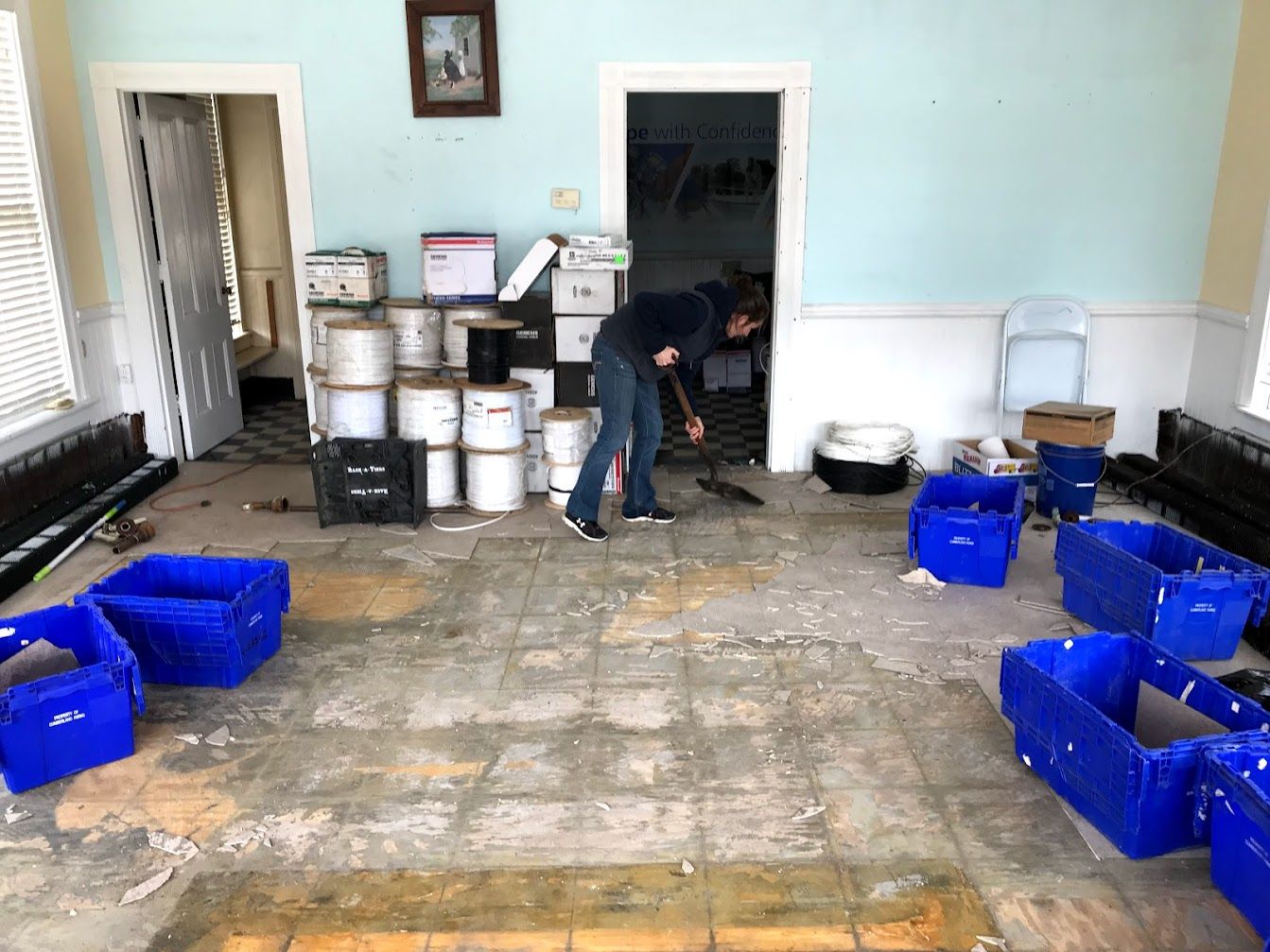 Person sweeping a room with blue bins, paint cans, and boxes. Old floor and light blue walls.
