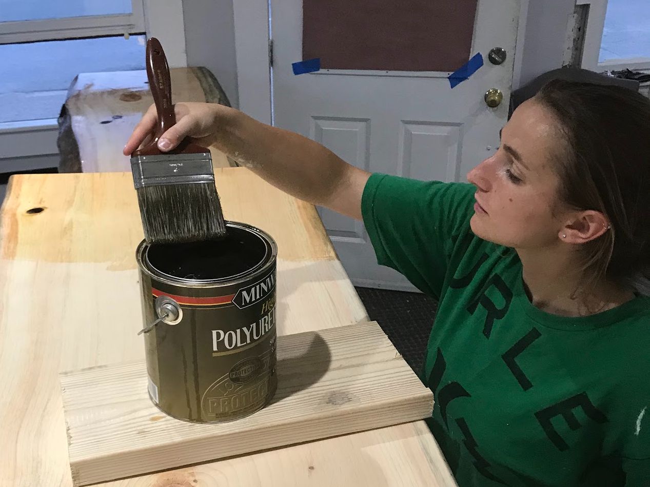 Woman dipping a paintbrush into a dark paint can on a wooden surface, preparing to paint.