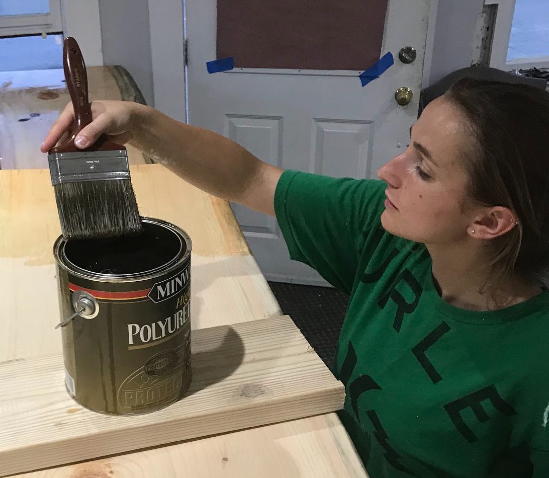 Woman dipping paintbrush into a can of stain on a wooden surface, preparing to apply finish.