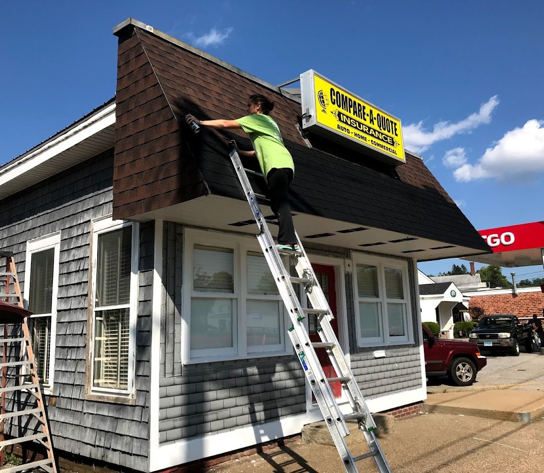 Person on ladder repairing roof of building with gray siding, next to a gas station.