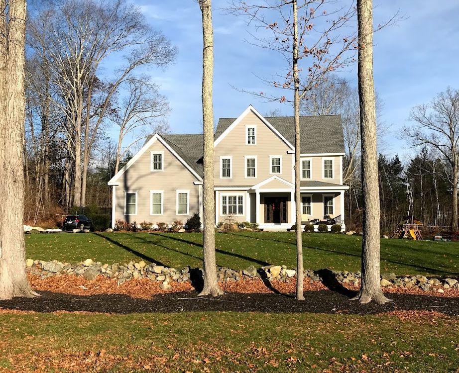 Two-story gray house with green lawn, trees in foreground, and blue sky.