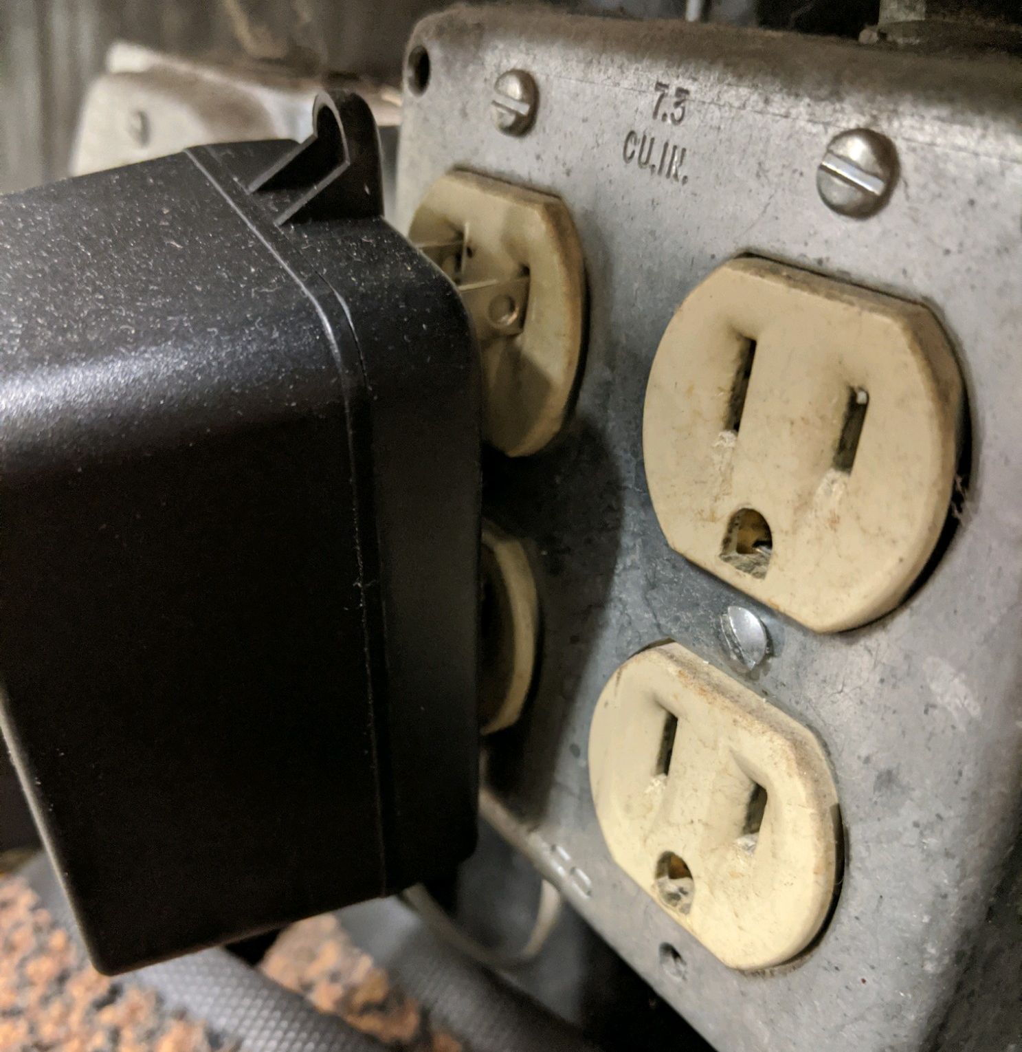 Close-up of a weathered metal electrical outlet box with two grounded outlets and a black plug inserted.