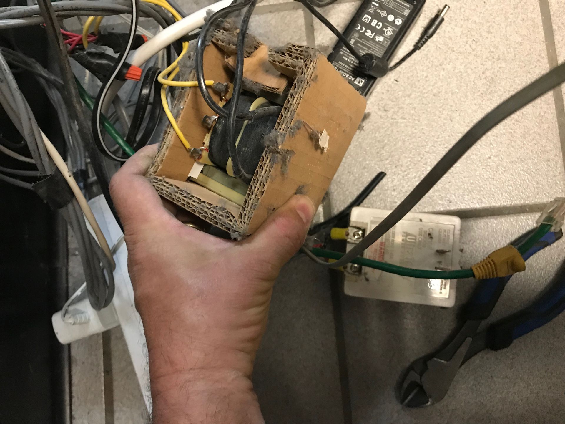 Hand holding a homemade electrical device in a cardboard box, wires and tools present near an outlet.