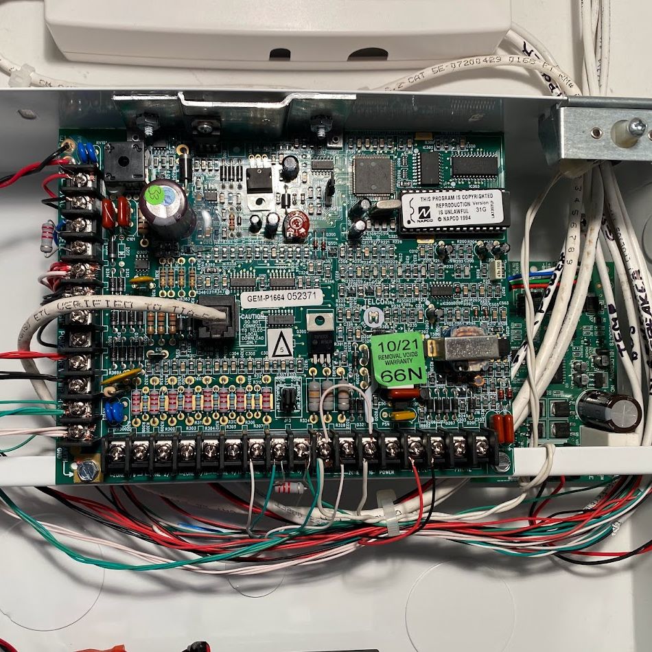 A circuit board in a white metal box. It has many wires and components. The board is green with black, red, and white wires.