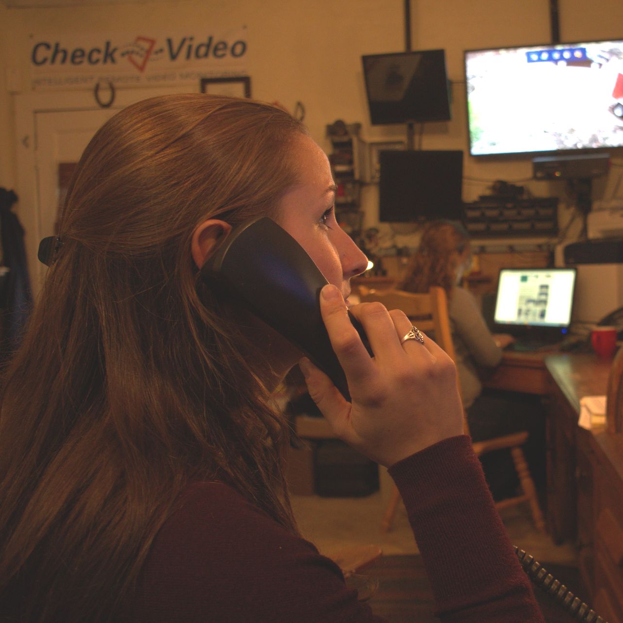 Woman with reddish-brown hair on a landline phone indoors, other person working on a computer in the background.