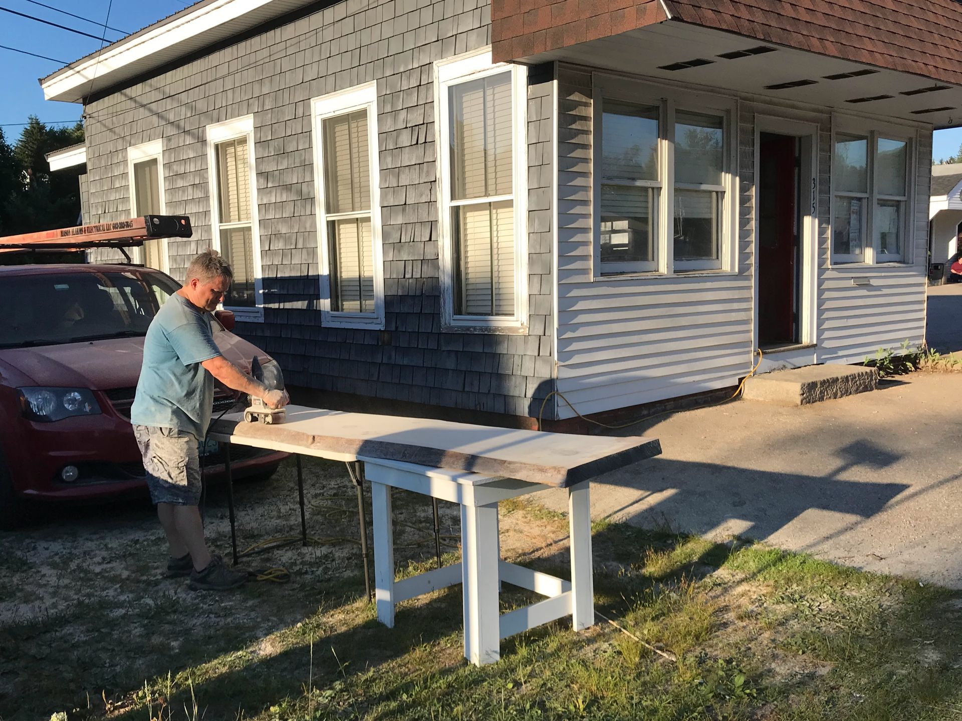Man sanding a wooden surface set on a white table outdoors, in front of a gray and white house.