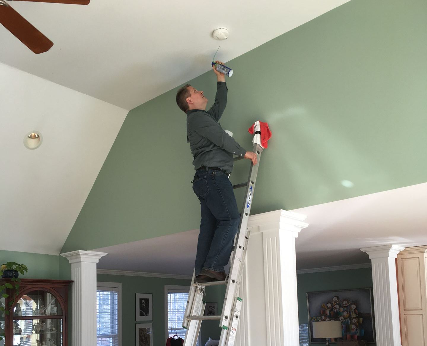 Man on a ladder installing a smoke detector on a high, green wall in a home.