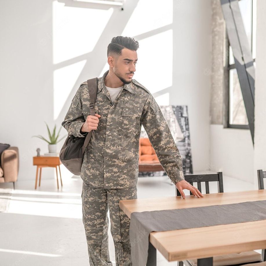 Man in camouflage uniform, carrying a bag, standing by a wooden table in a well-lit room.