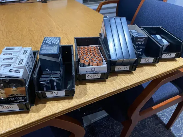 Several black storage bins filled with various items on a wooden table. Each bin has a label.