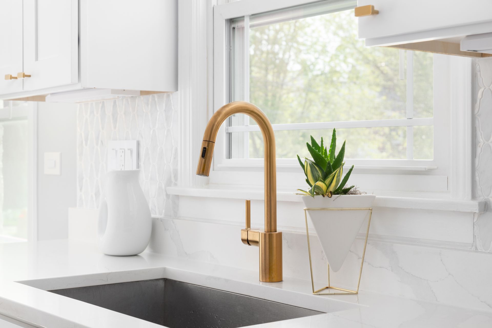 Gold faucet over a stainless steel sink in a bright white kitchen with a window and potted plant.