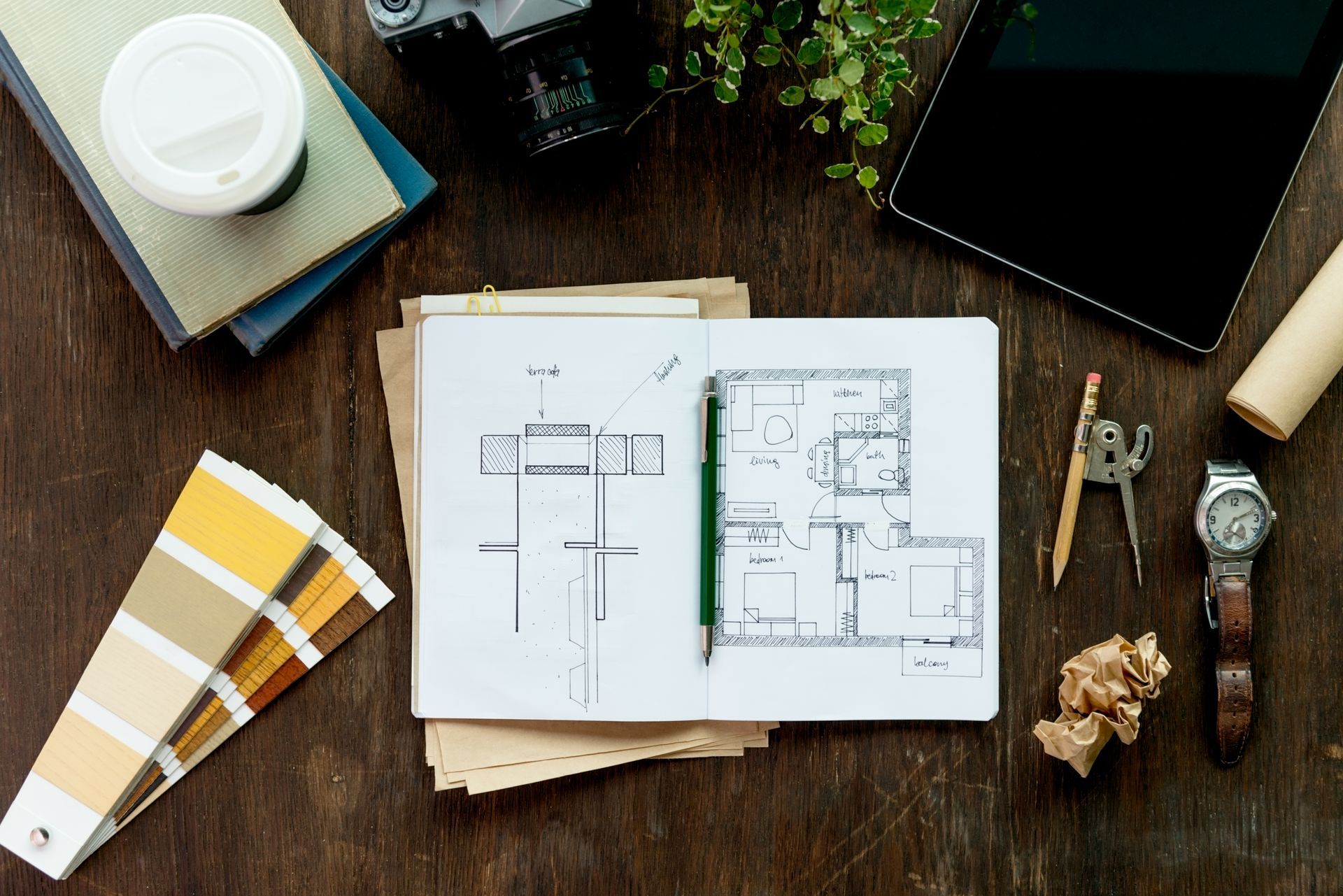 Overhead view of a design workspace; notebook with blueprints, color swatches, coffee cup, camera, and watch.
