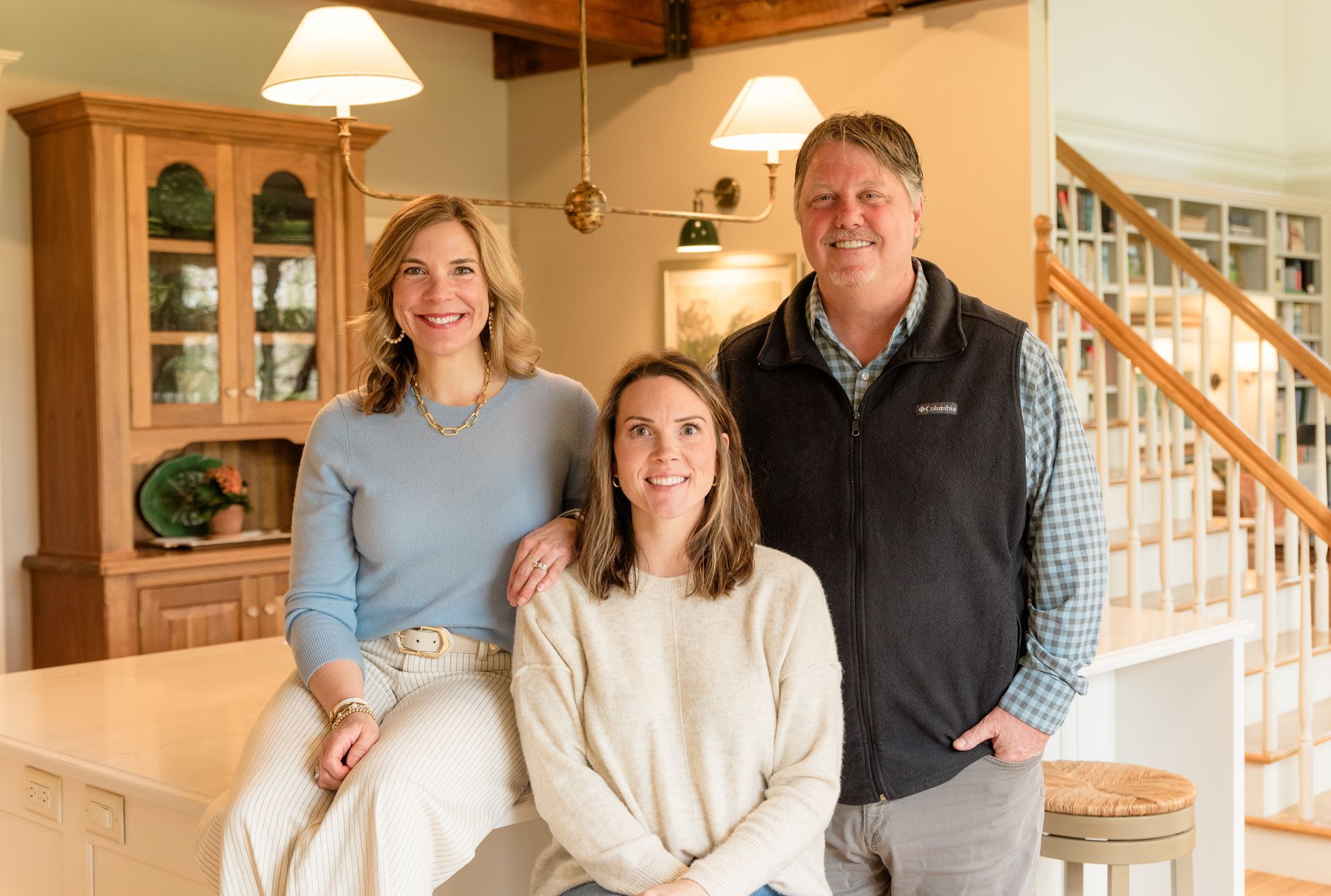 Three people pose in a kitchen: two women and a man. The women are seated; the man stands.