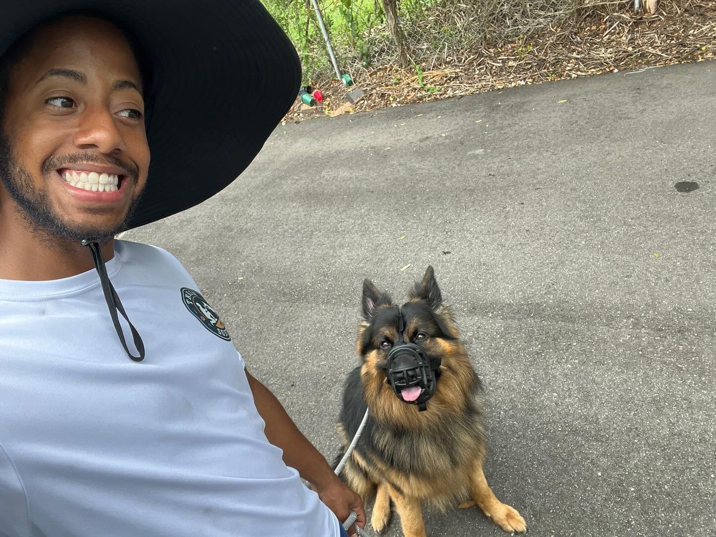 Smiling person with a wide-brim hat posing beside a fluffy dog on a paved path