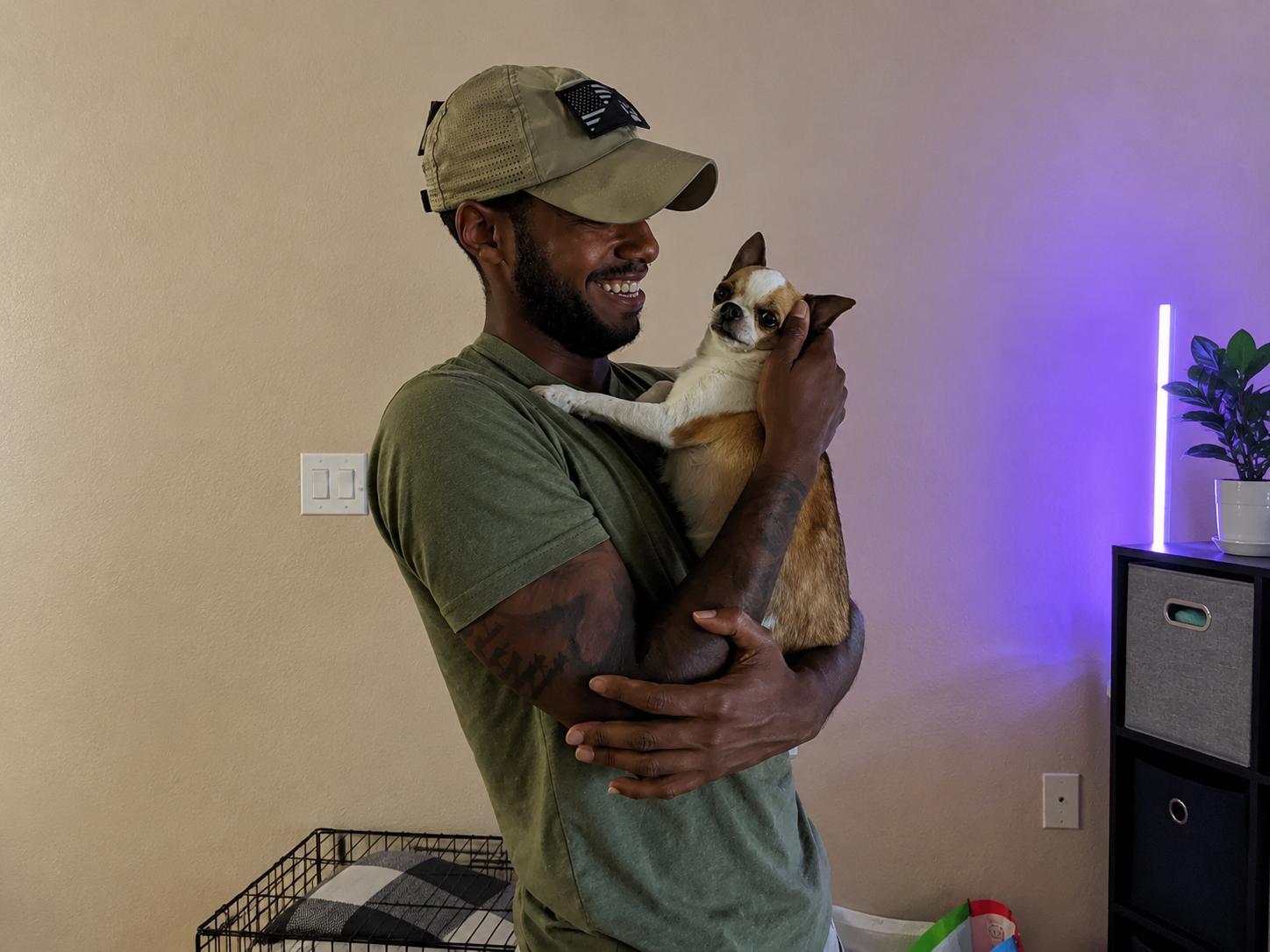 Person holding a small dog indoors, smiling near a beige wall with purple light and a plant in the background