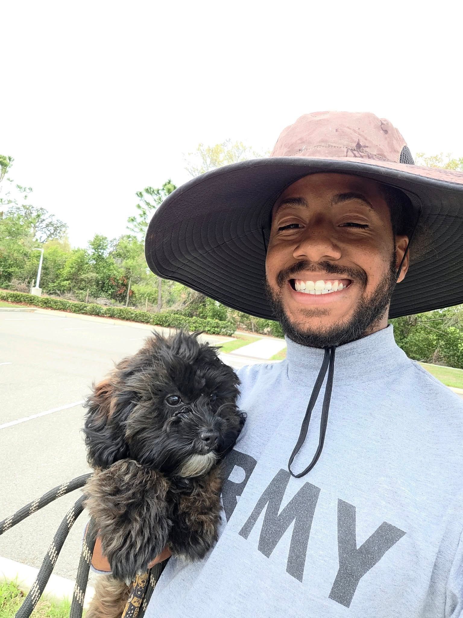 Smiling person in a wide-brim hat holding a small black dog outdoors