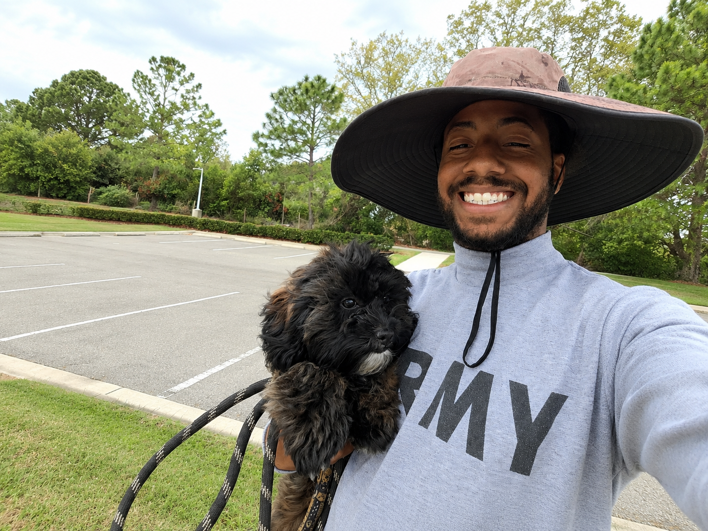 Smiling person in a wide-brim hat holding a black dog leash on a roadside trail with trees nearby