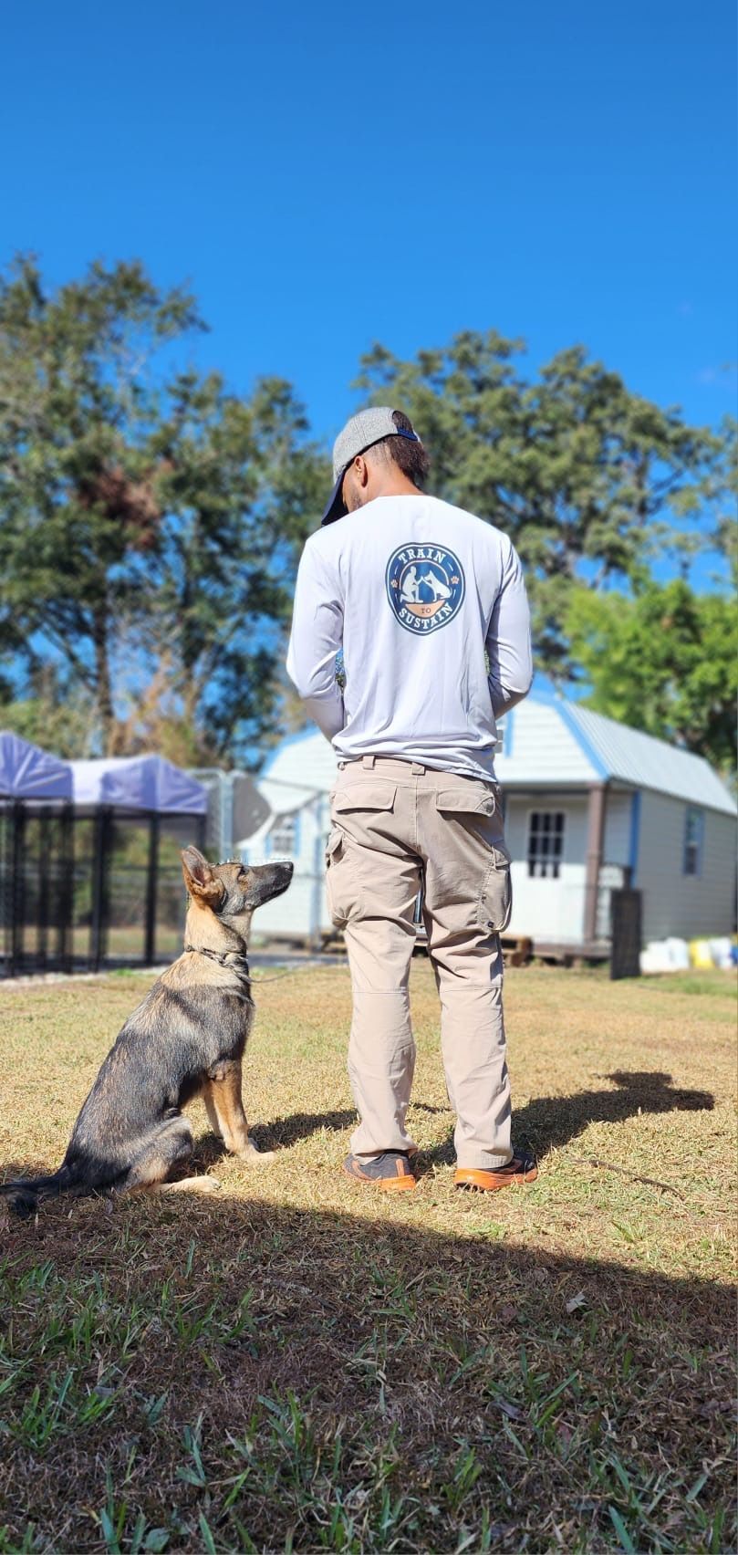 Person in white shirt and beige pants stands outdoors with a dog sitting attentively beside them.