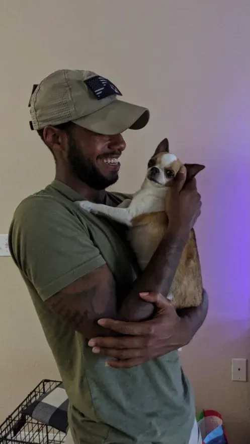 Man in green shirt and cap holding a small brown-and-white dog indoors