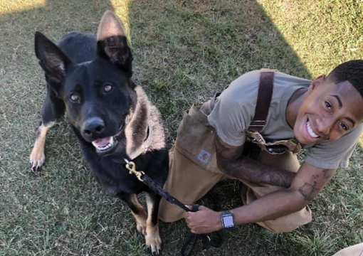 Person kneeling beside a black-and-tan dog on grass, both looking up at the camera.