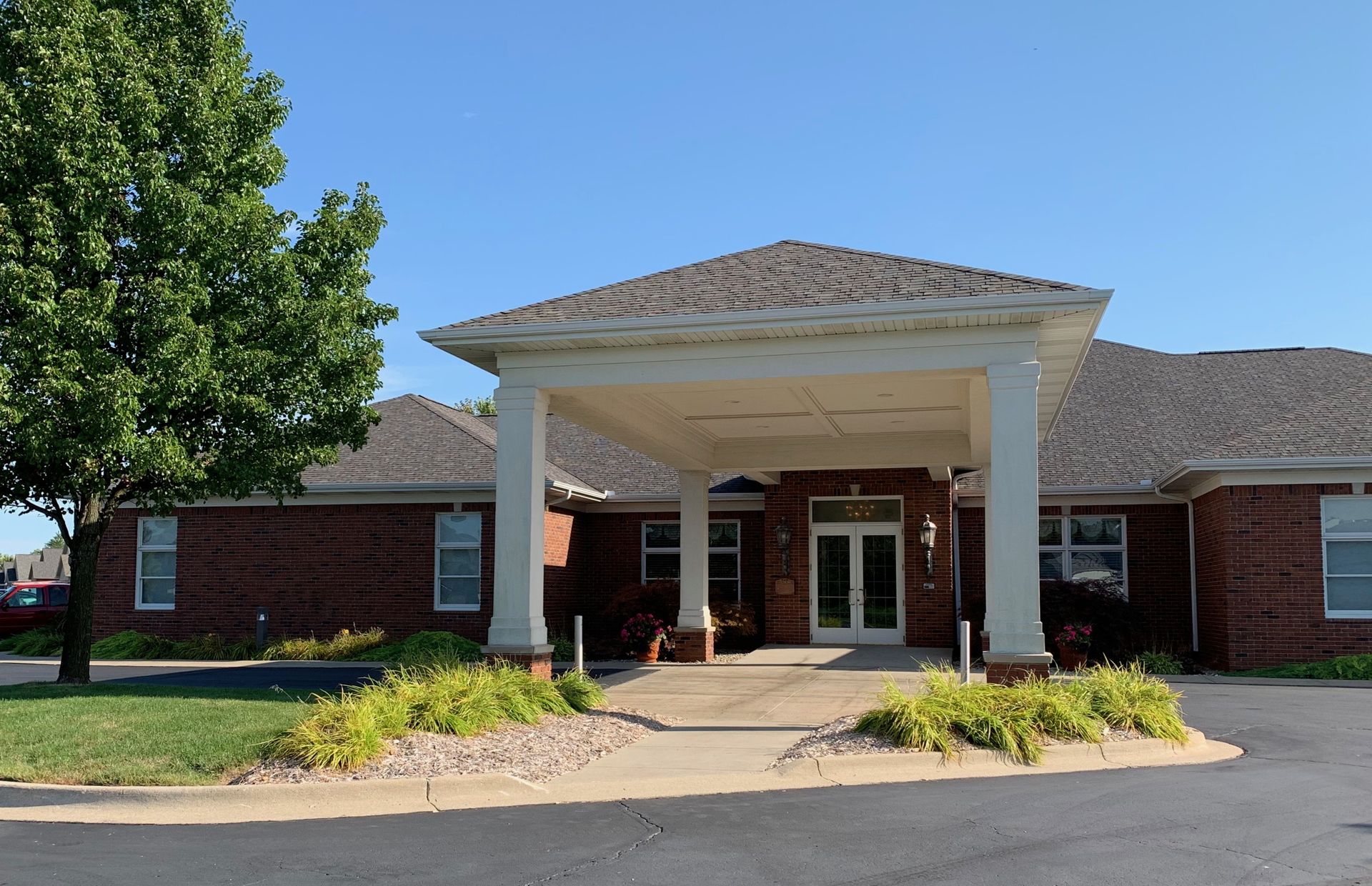 Front entrance of a brick building with white columns, a covered driveway, and landscaped shrubs under a clear sky