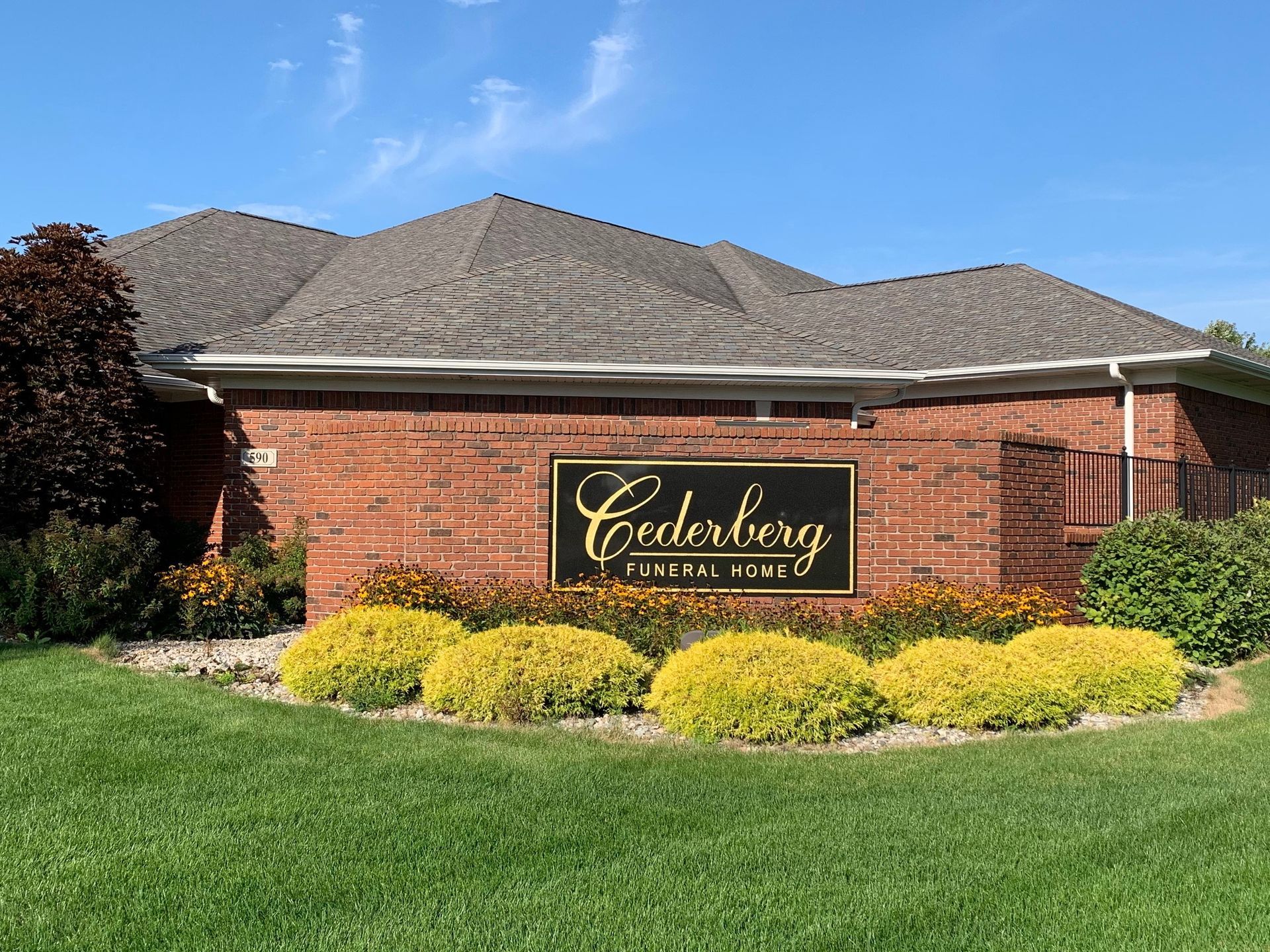 Brick entrance sign for Cedarsberg, with yellow shrubs and a green lawn in front of a building.