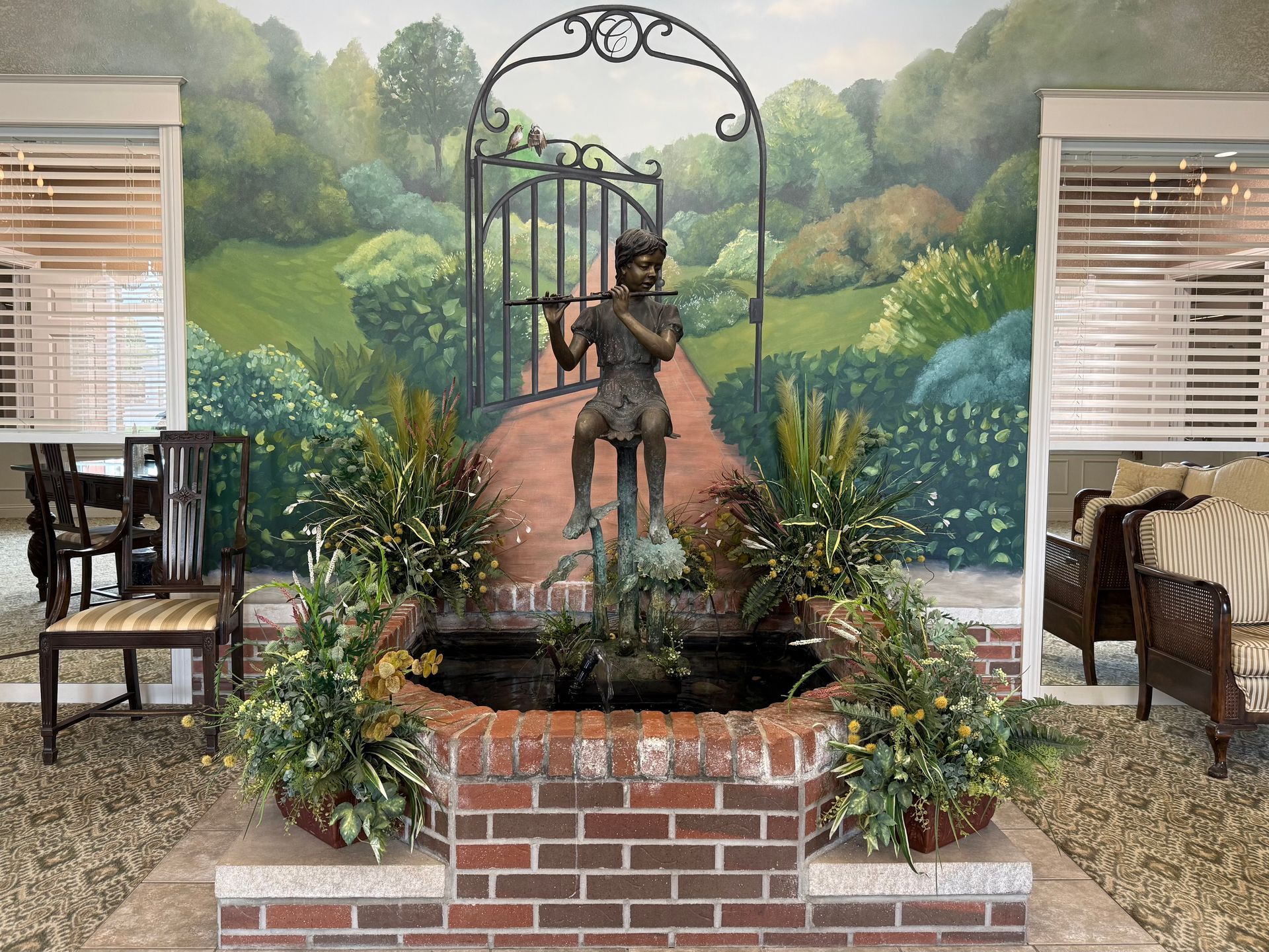 Garden fountain with bronze statue in front of a painted landscape mural, flanked by chairs and plants